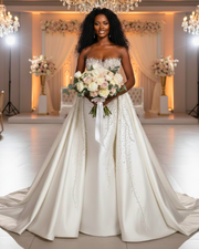 The dress features a fitted silhouette (likely mermaid or trumpet) with an elaborate overskirt. The sweetheart-neckline bodice and the entire front panel of the overskirt are densely hand-embellished with various sizes of clear crystals and white pearls, which appear to cascade downward from the bustline in a dripping effect. The fabric of the skirt and overskirt is a smooth, heavy satin.