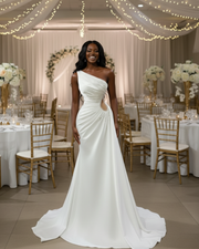 Modern one-shoulder satin wedding dress with an asymmetrical neckline, elegant draped bodice, beaded side cutout detail, and a sleek floor-length silhouette with a soft chapel-train.