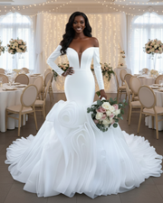 Statement off-the-shoulder mermaid wedding dress featuring a deep plunging neckline, fitted long sleeves, and an extravagant sculpted ruffle skirt with dramatic layered organza detailing in pure white.