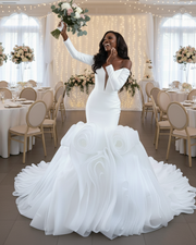 Statement off-the-shoulder mermaid wedding dress featuring a deep plunging neckline, fitted long sleeves, and an extravagant sculpted ruffle skirt with dramatic layered organza detailing in pure white.