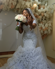 Luxurious embellished mermaid wedding dress featuring intricate silver beadwork, illusion neckline with crystal detailing, structured bodice with geometric patterns, detachable embellished cape, and a dramatic multi-layered ruffled skirt in bright white.