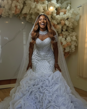 Luxurious embellished mermaid wedding dress featuring intricate silver beadwork, illusion neckline with crystal detailing, structured bodice with geometric patterns, detachable embellished cape, and a dramatic multi-layered ruffled skirt in bright white.