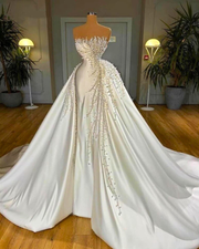 The dress features a fitted silhouette (likely mermaid or trumpet) with an elaborate overskirt. The sweetheart-neckline bodice and the entire front panel of the overskirt are densely hand-embellished with various sizes of clear crystals and white pearls, which appear to cascade downward from the bustline in a dripping effect. The fabric of the skirt and overskirt is a smooth, heavy satin.