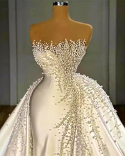 The dress features a fitted silhouette (likely mermaid or trumpet) with an elaborate overskirt. The sweetheart-neckline bodice and the entire front panel of the overskirt are densely hand-embellished with various sizes of clear crystals and white pearls, which appear to cascade downward from the bustline in a dripping effect. The fabric of the skirt and overskirt is a smooth, heavy satin.